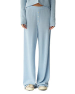 A person stands against a white background wearing Guest in Residence Everywear Cashmere Pant in light blue, featuring wide legs and a front button closure, paired with a matching long-sleeve top and white sneakers.