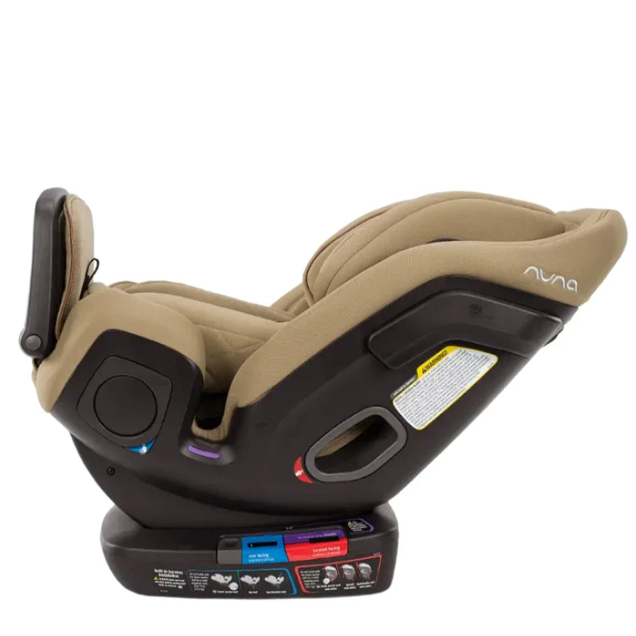 The Nuna EXEC with Slip Cover and 2nd Insert is a premium tan child car seat positioned rear-facing on a black base. It features adjustable straps, an Aeroflex side impact protection system, and safety instructions on the side. For added comfort and convenience, it includes a multi-functioning adjustable leg rest.