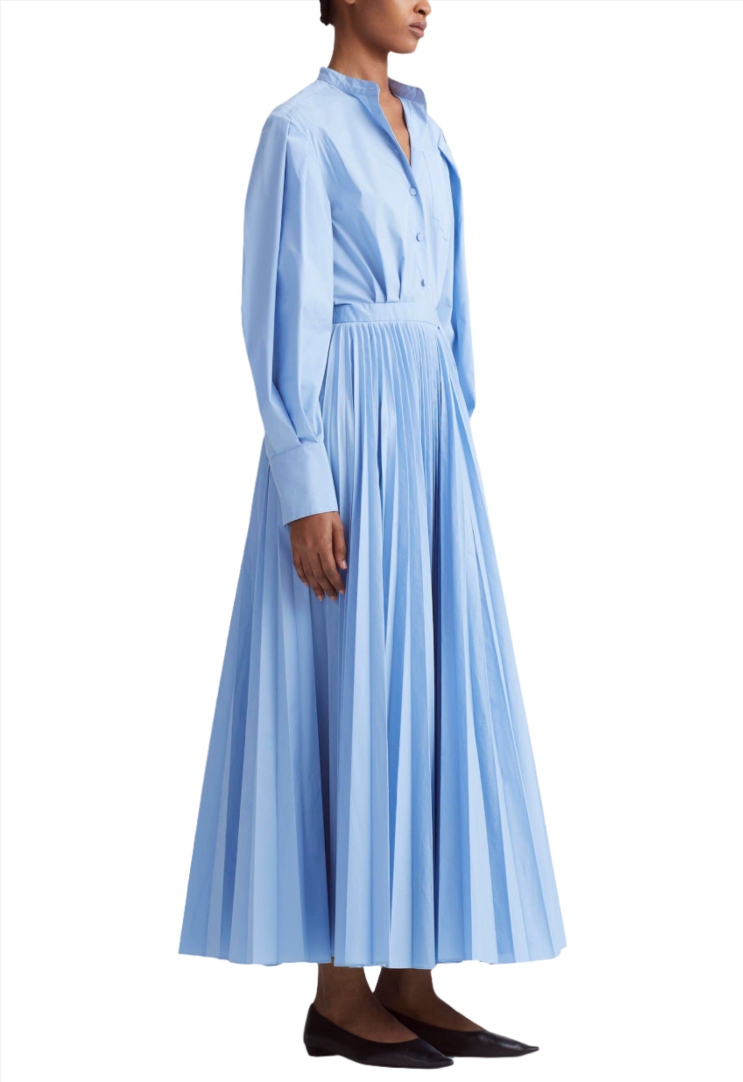 Person standing in an Altuzarra Binoche Dress by Altuzarra, a light blue, long-sleeved dress with balloon sleeves and an elasticated back, featuring a pleated skirt and paired with black pointed shoes, against a plain white background.