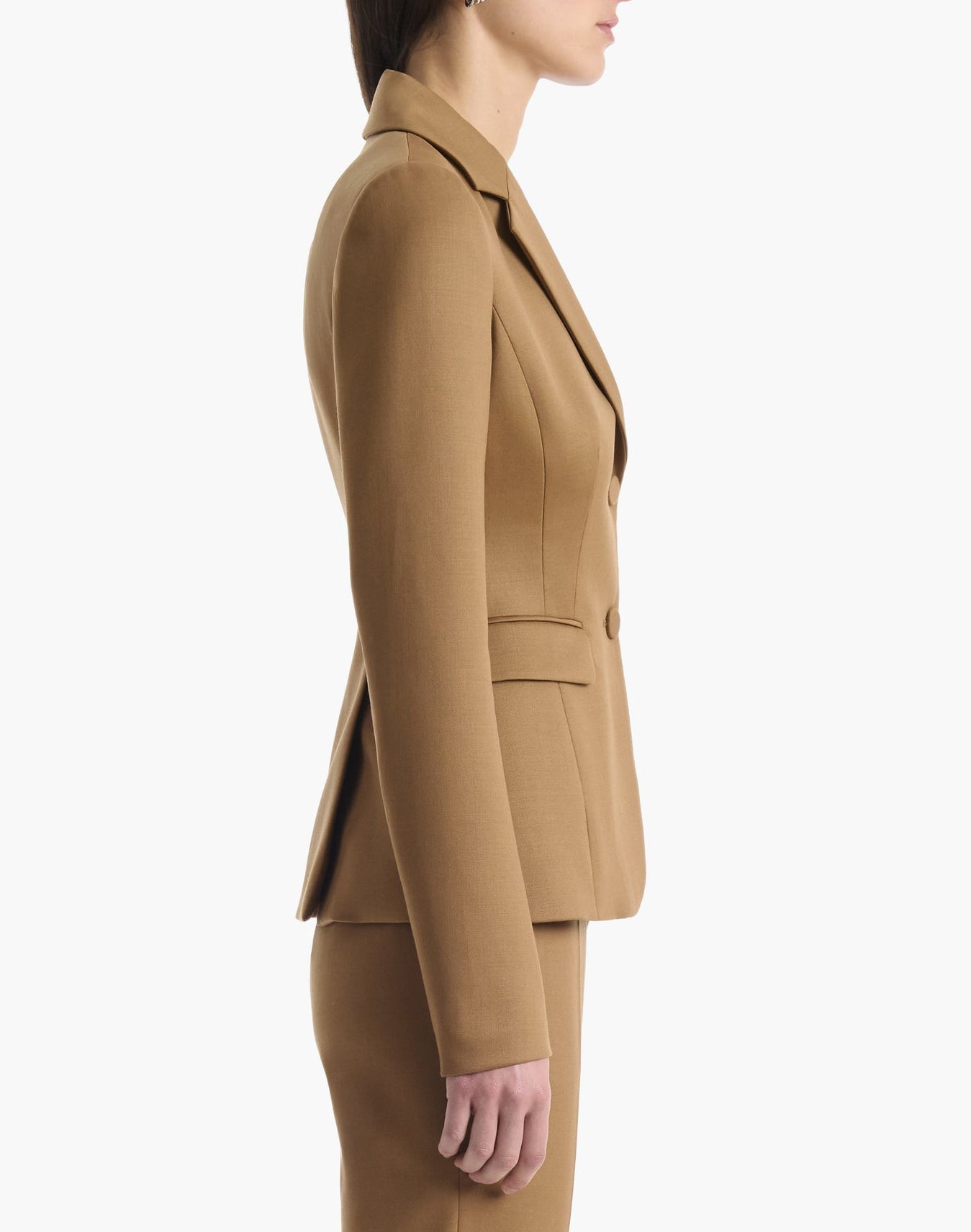 Clad in Altuzarra’s Fenice Jacket, this woman stands in profile against a white background, arms relaxed at her sides, effortlessly highlighting the impeccable suiting style of this tan virgin wool blend piece.