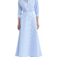Clad in the Altuzarra Eleanor Dress—a light blue, collared, button-down maxi dress crafted from cotton with three-quarter sleeves—a woman completes her look with black heeled sandals.