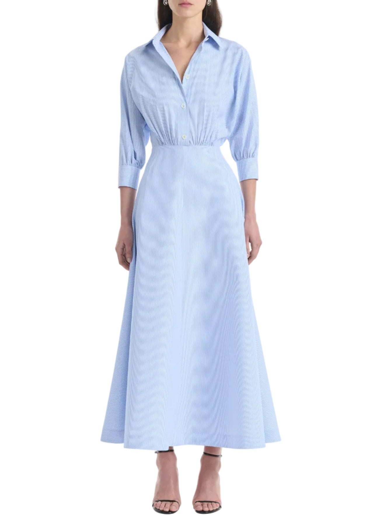 Clad in the Altuzarra Eleanor Dress—a light blue, collared, button-down maxi dress crafted from cotton with three-quarter sleeves—a woman completes her look with black heeled sandals.