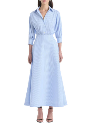 Clad in the Altuzarra Eleanor Dress—a light blue, collared, button-down maxi dress crafted from cotton with three-quarter sleeves—a woman completes her look with black heeled sandals.