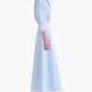 A woman stands in profile against a white background wearing the Eleanor Dress by Altuzarra—a light blue, long-sleeve, collared cotton dress made in Italy—paired with black high-heeled sandals.
