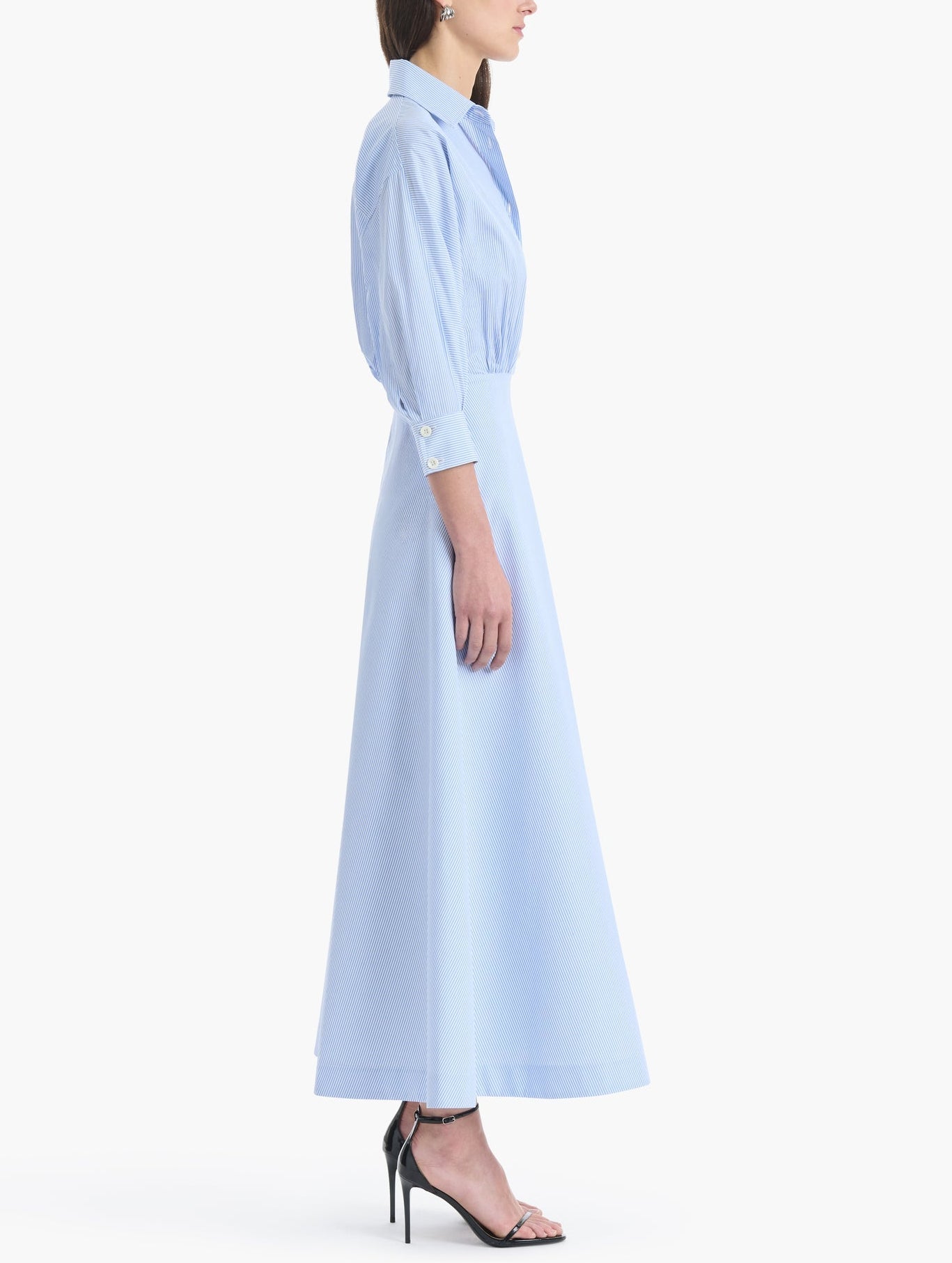 A woman stands in profile against a white background wearing the Eleanor Dress by Altuzarra—a light blue, long-sleeve, collared cotton dress made in Italy—paired with black high-heeled sandals.