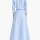 A woman stands with her back to the camera, wearing the Altuzarra Eleanor Dress—a light blue, long-sleeve, collared design featuring a flared skirt and paired with black high-heeled shoes. Expertly crafted in Italy from soft cotton, this dress captures an air of effortless elegance.