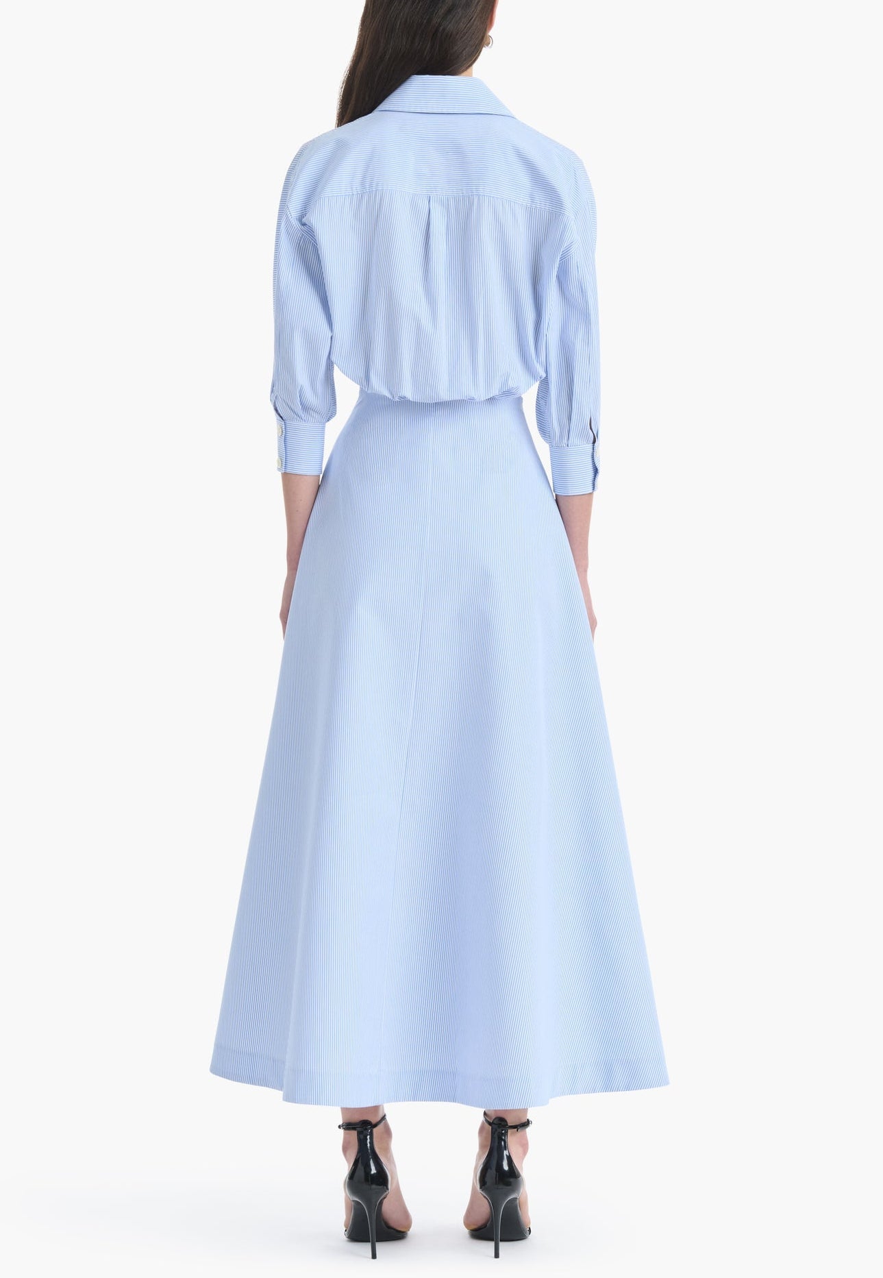 A woman stands with her back to the camera, wearing the Altuzarra Eleanor Dress—a light blue, long-sleeve, collared design featuring a flared skirt and paired with black high-heeled shoes. Expertly crafted in Italy from soft cotton, this dress captures an air of effortless elegance.