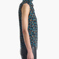 A woman stands in profile against a plain white background, wearing the Altuzarra Holt Top—a sleeveless, high-neck top with a blue and brown pattern and relaxed silhouette—paired with black pants.