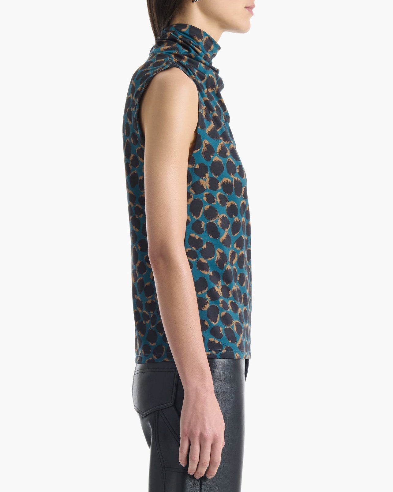 A woman stands in profile against a plain white background, wearing the Altuzarra Holt Top—a sleeveless, high-neck top with a blue and brown pattern and relaxed silhouette—paired with black pants.