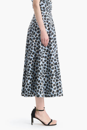 A woman stands in profile against a white background, wearing the Altuzarra Varda Skirt—a blue and black leopard print A-line midi skirt—paired with black high-heeled sandals.