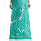 A person wearing the Altuzarra Linnie Silk Skirt—a long green silk skirt with a straight silhouette and fitted waist, adorned with a white and black fishnet and fish pattern—stands in black sandals against a white background.