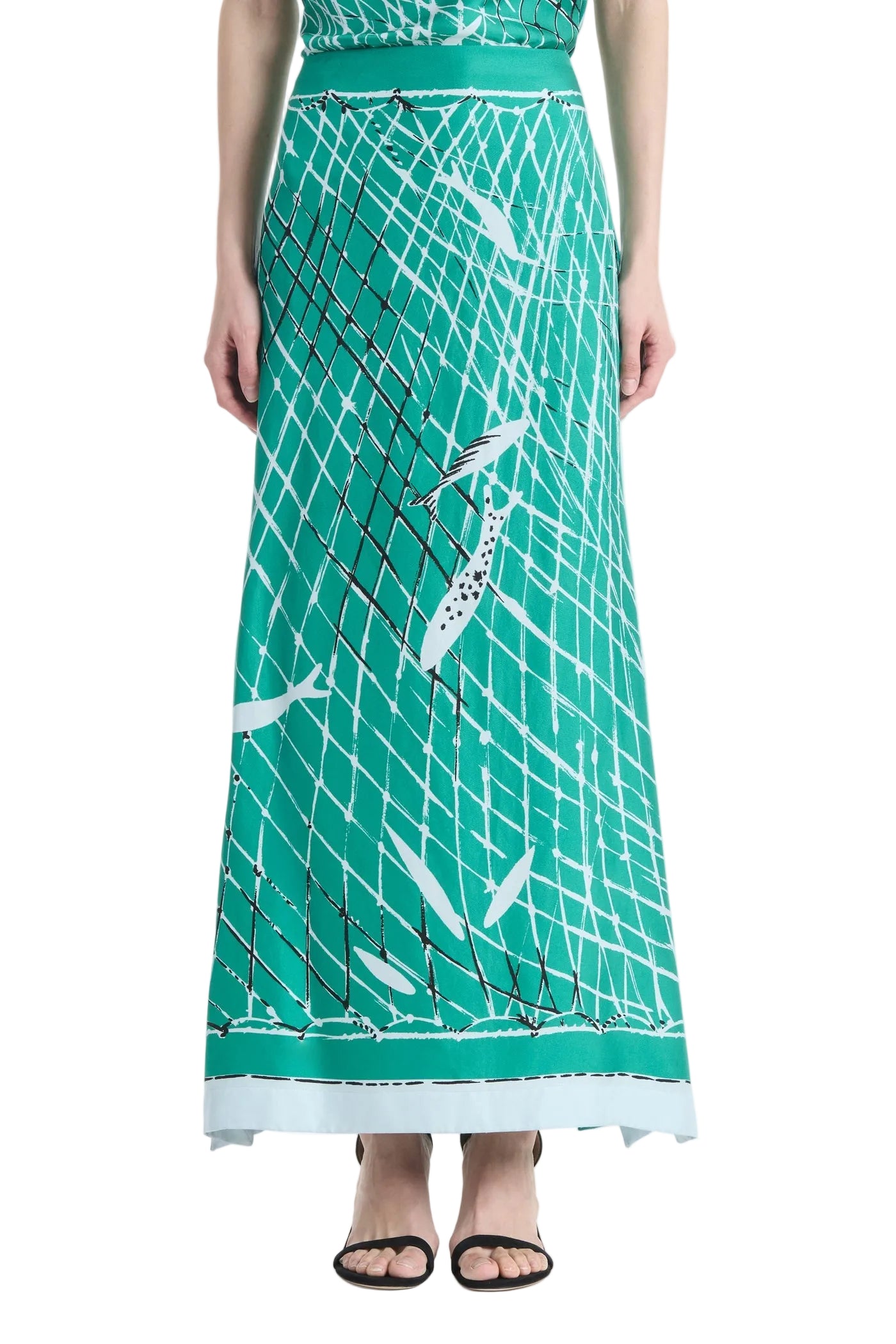 A person wearing the Altuzarra Linnie Silk Skirt—a long green silk skirt with a straight silhouette and fitted waist, adorned with a white and black fishnet and fish pattern—stands in black sandals against a white background.