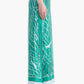 A person stands in profile against a white background wearing teal wide-leg pants with a white and black geometric pattern and black open-toe heeled sandals, channeling the modern elegance of the Linnie Silk Skirt by Altuzarra.