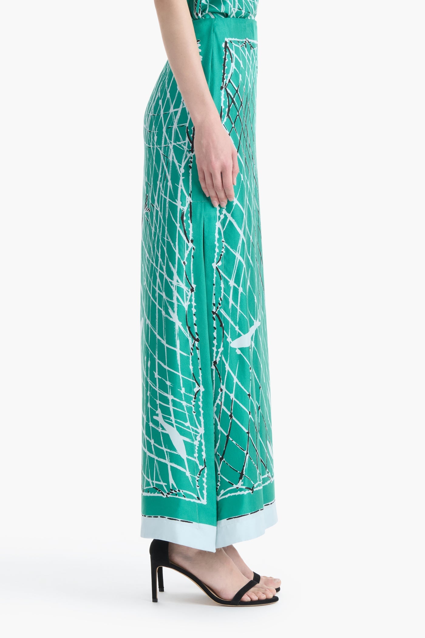 A person stands in profile against a white background wearing teal wide-leg pants with a white and black geometric pattern and black open-toe heeled sandals, channeling the modern elegance of the Linnie Silk Skirt by Altuzarra.