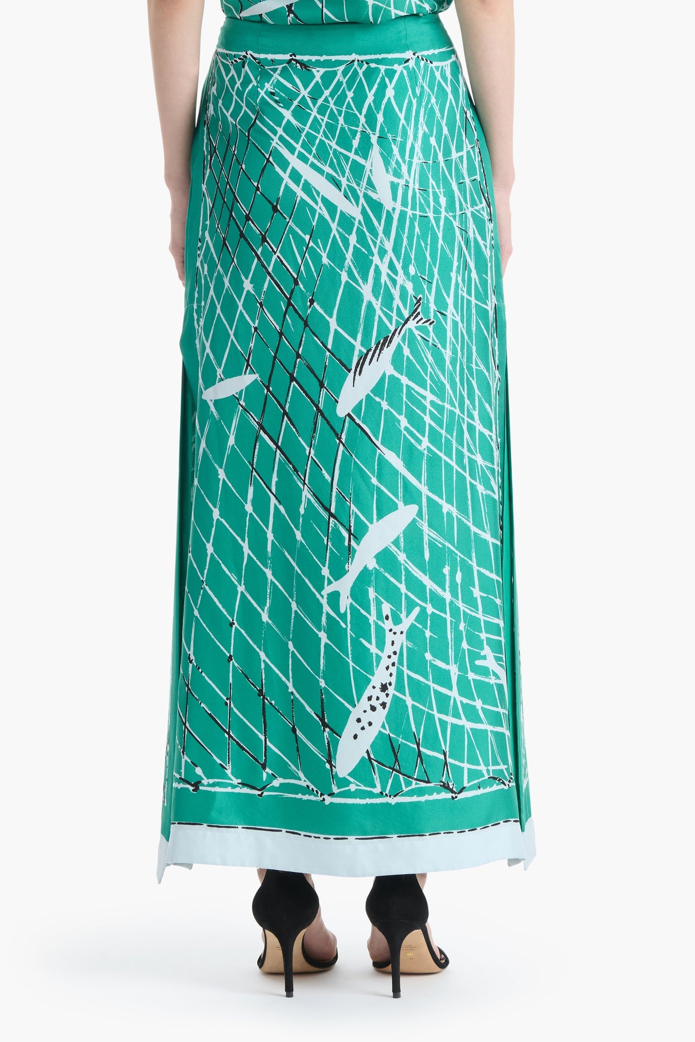 A person wearing the Altuzarra Linnie Silk Skirt, shown from the back, styled with black high-heeled sandals. The skirt features a green fish and net pattern, a fitted waist, and a straight silhouette for an elegant look.
