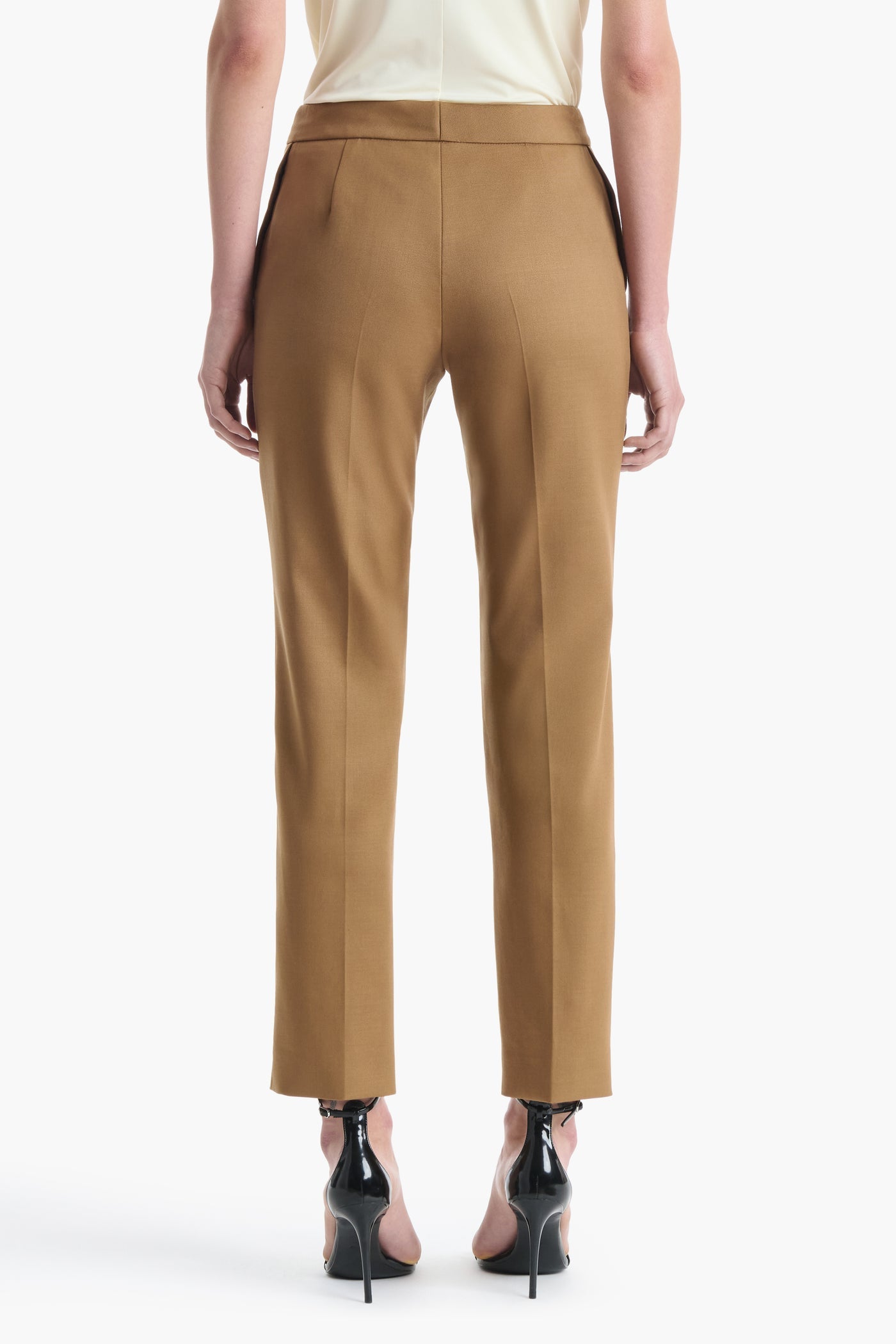 A person wearing the Altuzarra Todd Pant—tailored tan dress pants with a straight leg—paired with black high heels, shown from the back against a white background.