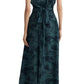 A person is standing with their back to the camera, wearing a sleeveless, dark green patterned Altuzarra Penny dress with a tied waist and black shoes. The elegant maxi dress flows beautifully, showcasing the exquisite design made in Italy by Altuzarra.