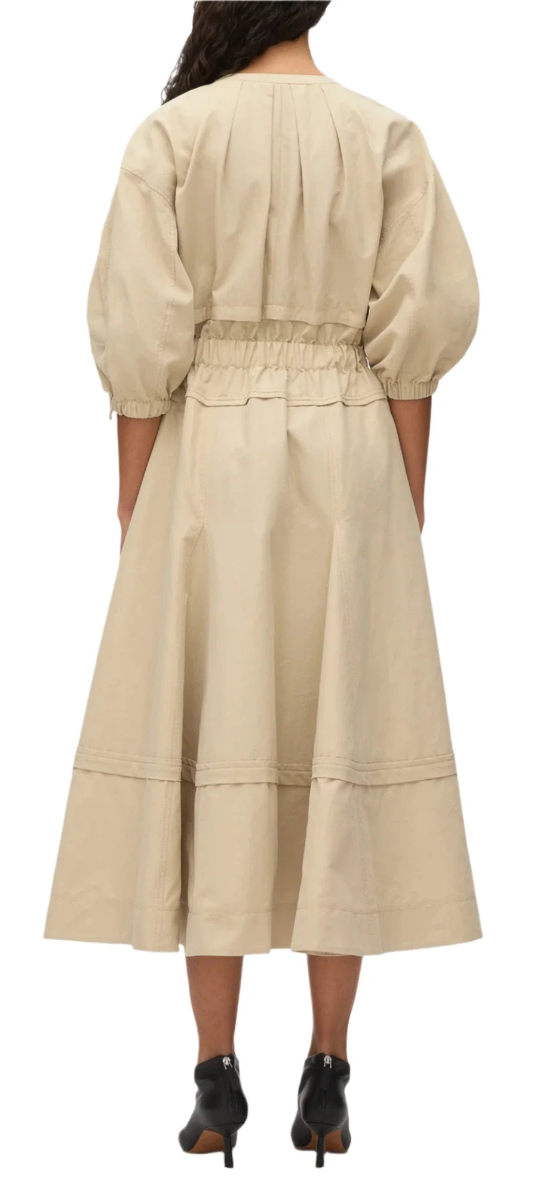 A person is seen wearing the 3.1 Phillip Lim V Neck Dress with Gathered Sleeves, a beige, long-sleeved dress that extends to the ankle and features belle sleeves and a gathered waist. The dress is crafted from Italian ottoman cotton, with the individual standing back to the camera.