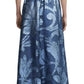A person is standing with their back facing the camera, wearing a Lela Rose Metallic Floral Fil Coupe Gown. The gown features a long, blue design adorned with baroque damask botanicals, a sheer top, a tied waist, and a full skirt that flows gracefully.