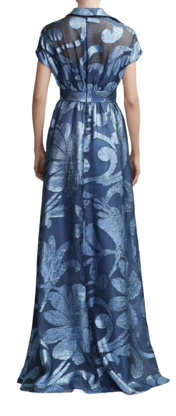 A person is standing with their back facing the camera, wearing a Lela Rose Metallic Floral Fil Coupe Gown. The gown features a long, blue design adorned with baroque damask botanicals, a sheer top, a tied waist, and a full skirt that flows gracefully.