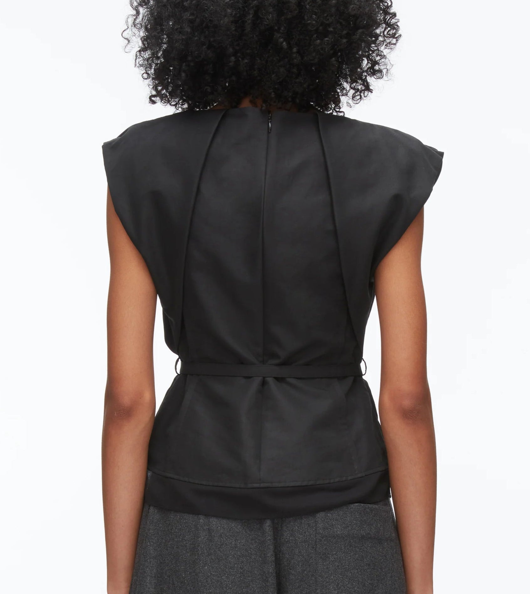 A person with curly hair is shown from the back, wearing the 3.1 Phillip Lim Sleeveless Draped Bow Top w Belt in black, featuring a sculptural bow neckline and fitted waist, paired with dark gray pants.
