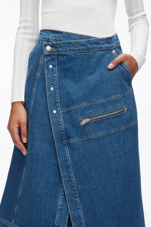 A person wears a white ribbed long-sleeve top and the 3.1 Phillip Lim Denim Wrap Skirt, a blue asymmetric midi skirt with front buttons and a zippered pocket.
