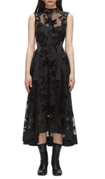 A woman in the 3.1 Phillip Lim Flocked Organza Sleeveless Midi Dress Combo, featuring a belted waist and sheer floral overlay, pairs it with black lace-up ankle boots for a modern occasion look against a white background.
