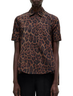 A model wears the Adam Lippes Trapeze Shirt Printed Poplin Leopard—a short-sleeve, collared top in brown and black leopard print cotton poplin—paired with black pants, front view against a white background.