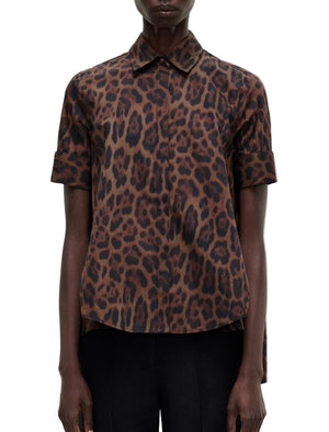 A model wears the Adam Lippes Trapeze Shirt Printed Poplin Leopard—a short-sleeve, collared top in brown and black leopard print cotton poplin—paired with black pants, front view against a white background.