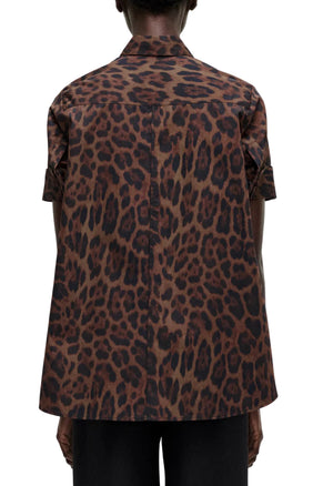 Person seen from the back wearing the Adam Lippes Trapeze Shirt Printed Poplin Leopard, a short-sleeve brown leopard print shirt in Italian cotton poplin, with black pants visible below the hem.