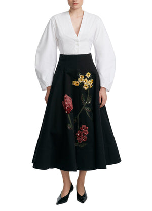 A person wearing a Lela Rose Button Front Peplum Top with a tailored waist and a black A-line skirt with colorful floral embroidery, paired with black pointed-toe shoes.