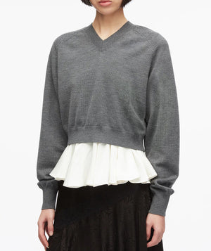 A woman wearing the 3.1 Phillip Lim Ultrafine Merino V Neck Pullover in grey, styled over a white ruffled blouse and paired with a black skirt, stands against a plain white background.