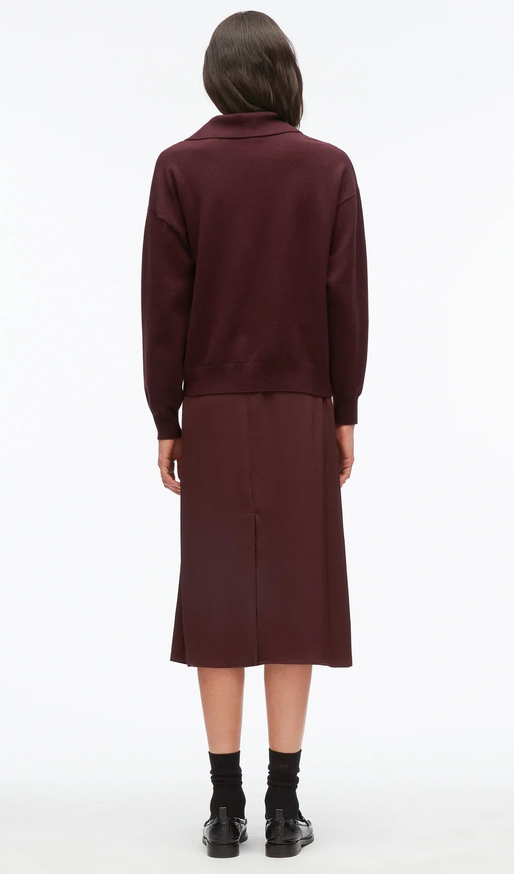 A person stands facing away, wearing the Ultrafine Merino Polo Midi Dress by 3.1 Phillip Lim in burgundy, paired with black socks and black shoes, against a plain white background.