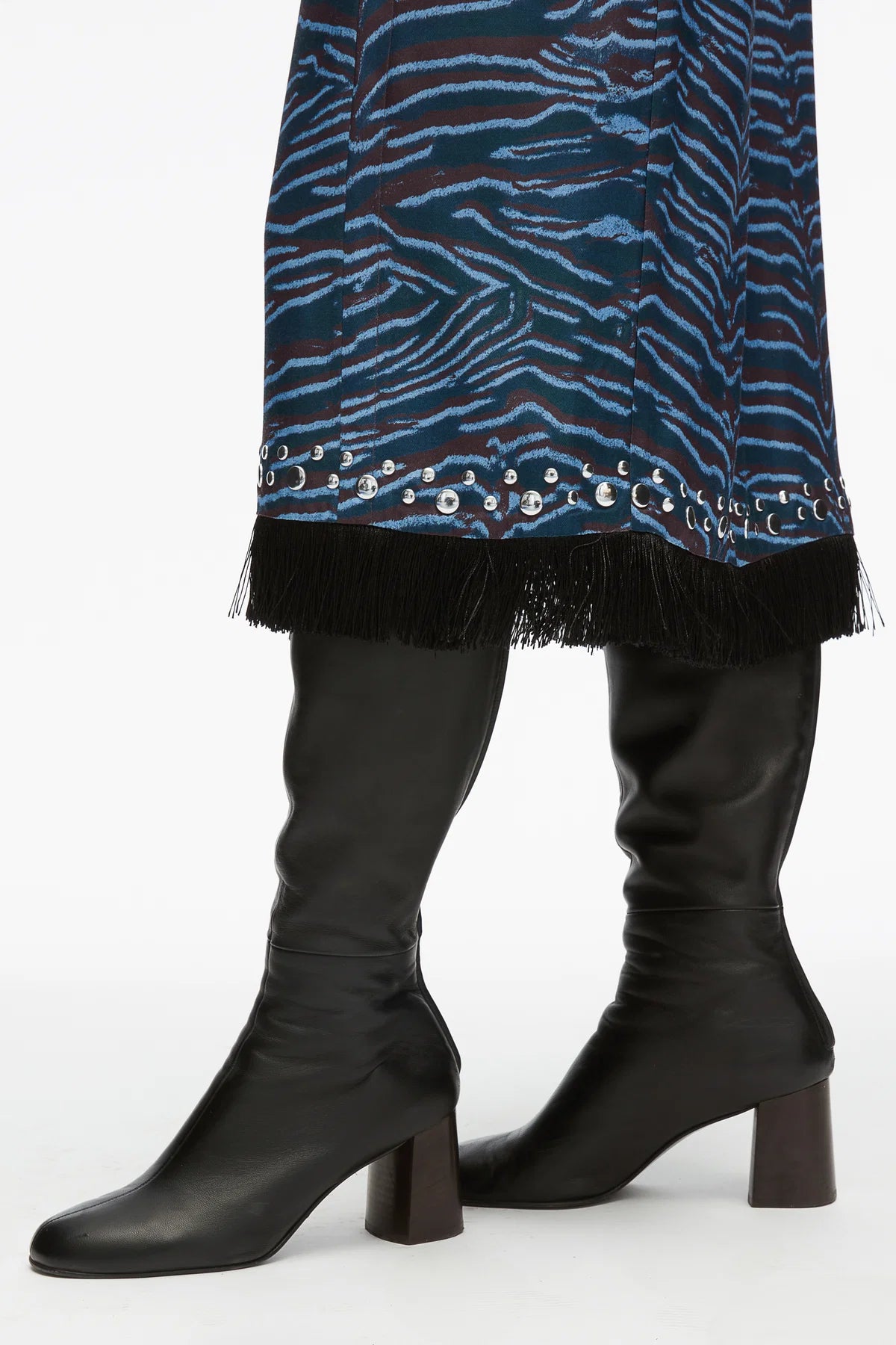 A person wearing the 3.1 Phillip Lim Tiger Print Studded Midi Dress, featuring a blue pattern with a fringed hem and silver studs, paired with black knee-high heeled leather boots.
