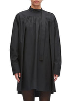 A person is wearing the 3.1 Phillip Lim Long Sleeve Poplin Mini Dress—a loose-fitting black cotton poplin design with a high neck, attached necktie detail, and a gathered silhouette—while standing against a plain background.