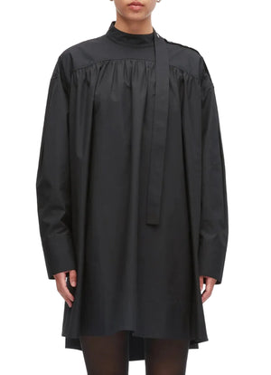 A person is wearing the 3.1 Phillip Lim Long Sleeve Poplin Mini Dress—a loose-fitting black cotton poplin design with a high neck, attached necktie detail, and a gathered silhouette—while standing against a plain background.