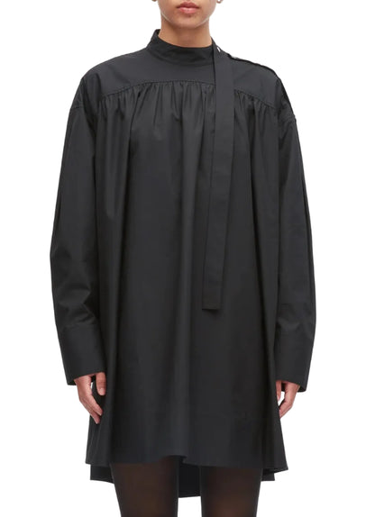 A person is wearing the 3.1 Phillip Lim Long Sleeve Poplin Mini Dress—a loose-fitting black cotton poplin design with a high neck, attached necktie detail, and a gathered silhouette—while standing against a plain background.