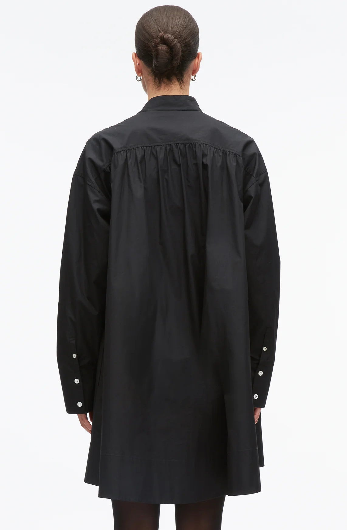A person wearing the 3.1 Phillip Lim Long Sleeve Poplin Mini Dress, featuring a loose-fitting black silhouette with gathered detailing, is shown from the back with hair in a bun and silver hoop earrings.