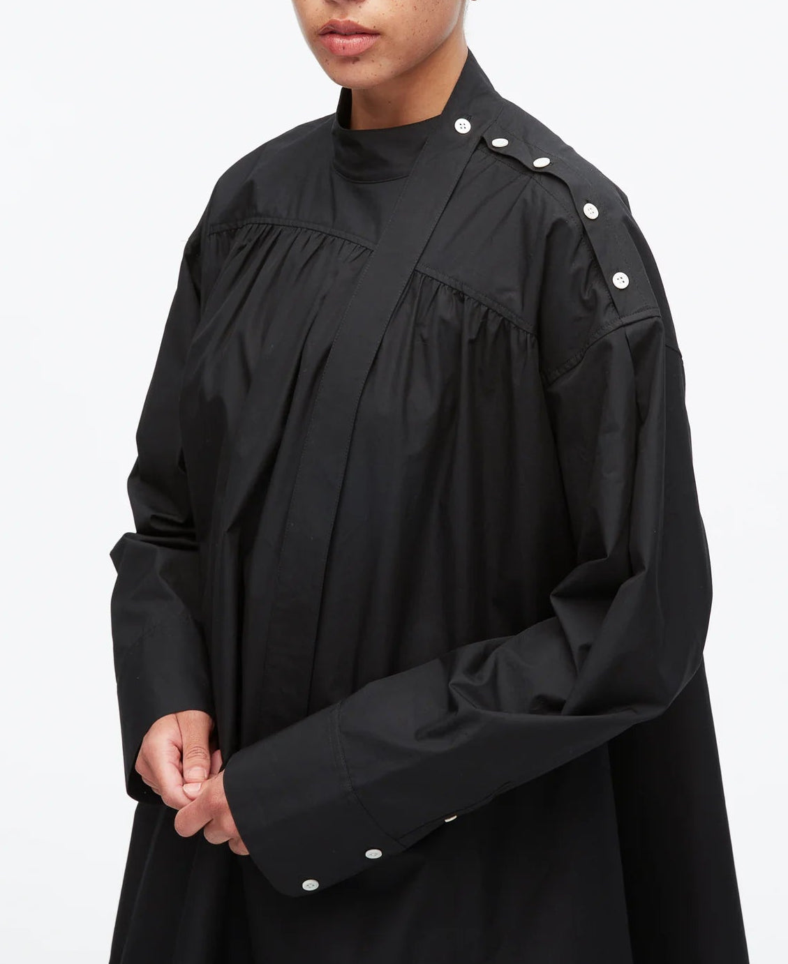A person wearing the 3.1 Phillip Lim Long Sleeve Poplin Mini Dress, a loose-fitting black dress with white snap buttons on the shoulder and cuff and utilitarian detailing, stands against a white background.