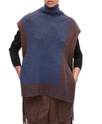 A person wears the 3.1 Phillip Lim Bicolor Rib Funnel Neck Vest—an oversized, sleeveless turtleneck sweater in blue and brown alpaca blend with painterly color blocking—layered over a black shirt and brown pants, standing with hands in pockets.
