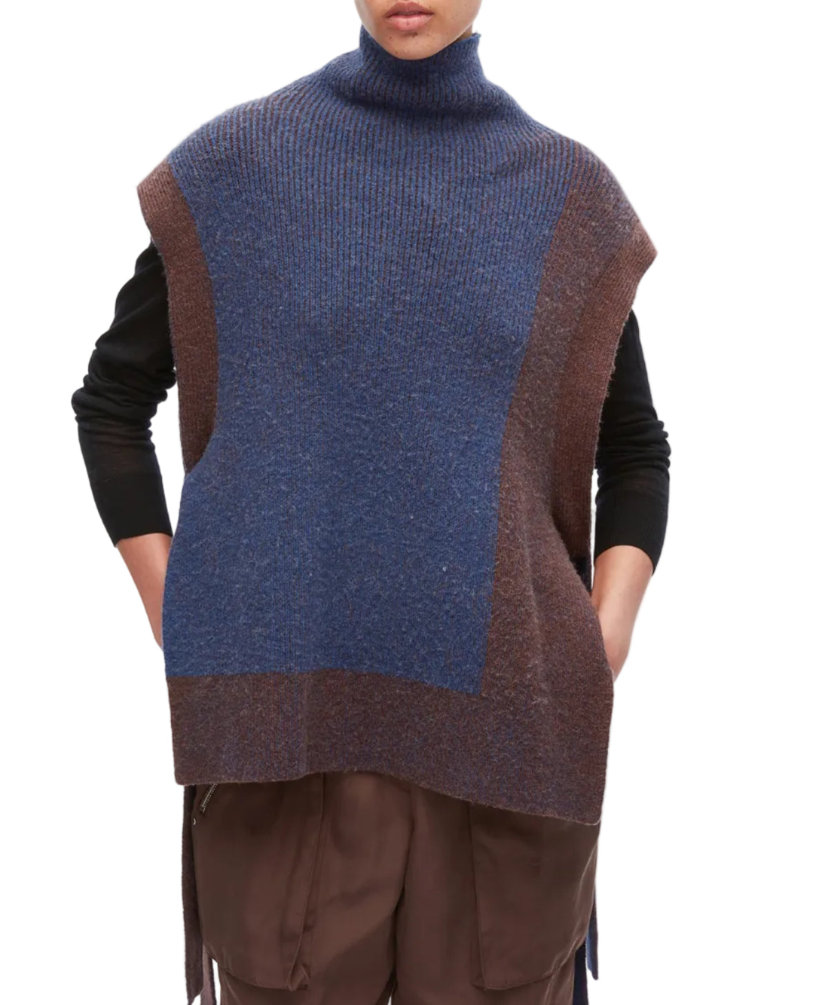 A person wears the 3.1 Phillip Lim Bicolor Rib Funnel Neck Vest—an oversized, sleeveless turtleneck sweater in blue and brown alpaca blend with painterly color blocking—layered over a black shirt and brown pants, standing with hands in pockets.