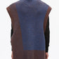 A person with dark hair in a bun is shown from the back against a plain light background, wearing the 3.1 Phillip Lim Bicolor Rib Funnel Neck Vest—an oversized sleeveless knit vest featuring painterly blue and brown color blocking—layered over a black top and brown pants.
