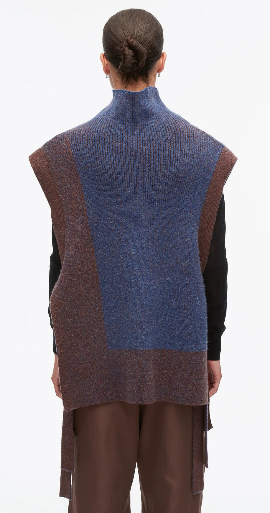 A person with dark hair in a bun is shown from the back against a plain light background, wearing the 3.1 Phillip Lim Bicolor Rib Funnel Neck Vest—an oversized sleeveless knit vest featuring painterly blue and brown color blocking—layered over a black top and brown pants.