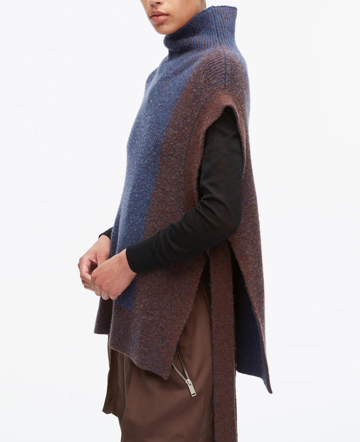 A person wears the 3.1 Phillip Lim Bicolor Rib Funnel Neck Vest—a sleeveless, oversized knit in painterly blue and brown alpaca blend—layered over a black shirt and brown zippered pants, standing against a white background.