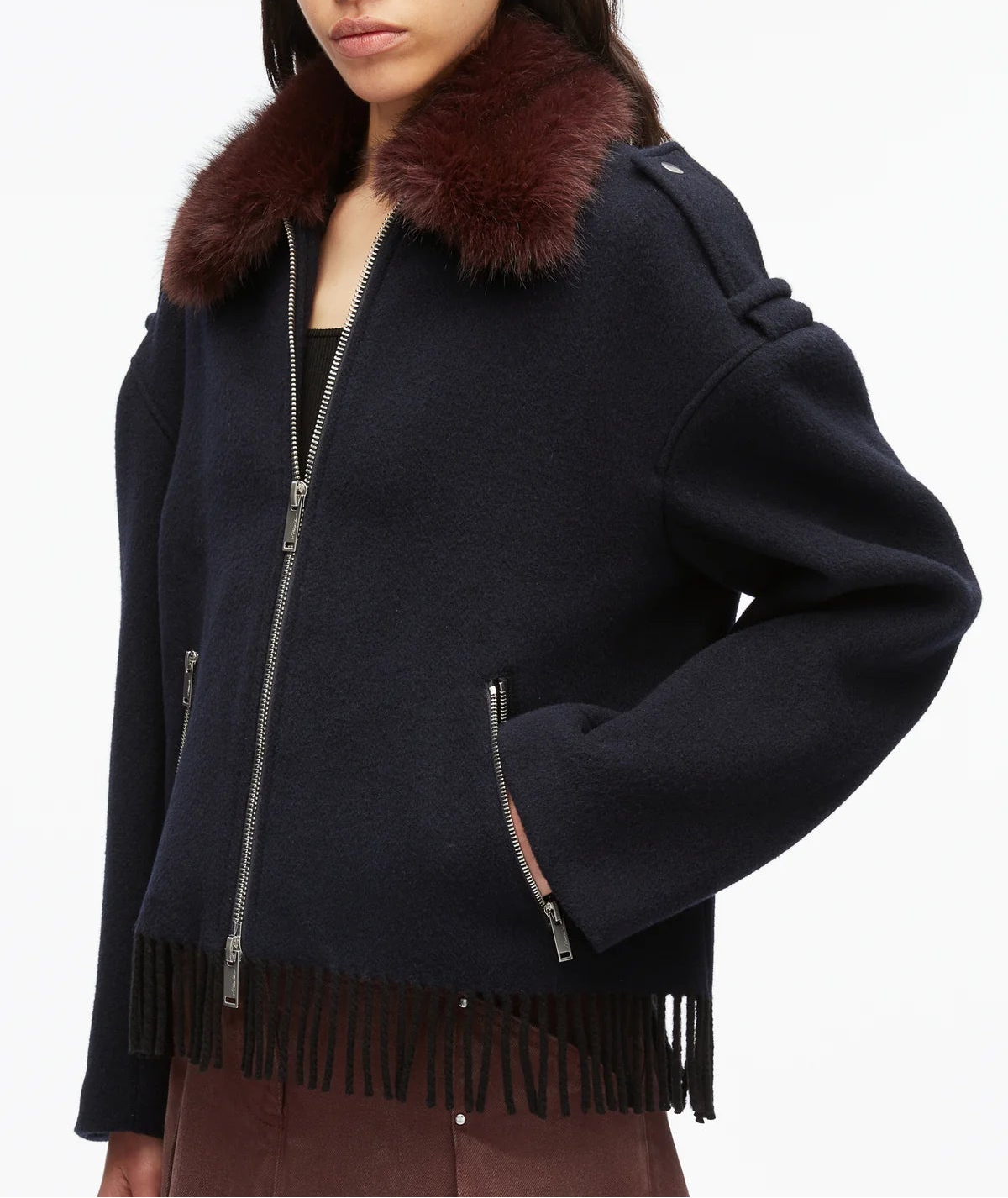 A person wears the 3.1 Phillip Lim Wool Blanket Zip Up Jacket—a dark handcrafted outerwear piece featuring a brown faux fur collar and fringe at the hem—paired with brown pants.