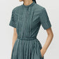 A person wears the Doron Stripe Shirting Dress by Adam Lippes—green with white vertical stripes, short sleeves, belted waist, mandarin collar, hands in pockets.