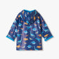 The Hatley Boys’ Colourful Dinos Rain Coat features a blue background with vibrant dinosaur prints, shown from the back with a hood and rolled cuffs.