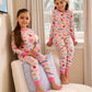 Two young girls wear matching Hatley Girls’ Delicious Candies Bamboo Pajama Sets as they pose in a sunlit room by the window—one sitting on a chair holding flowers, the other perched on the windowsill.