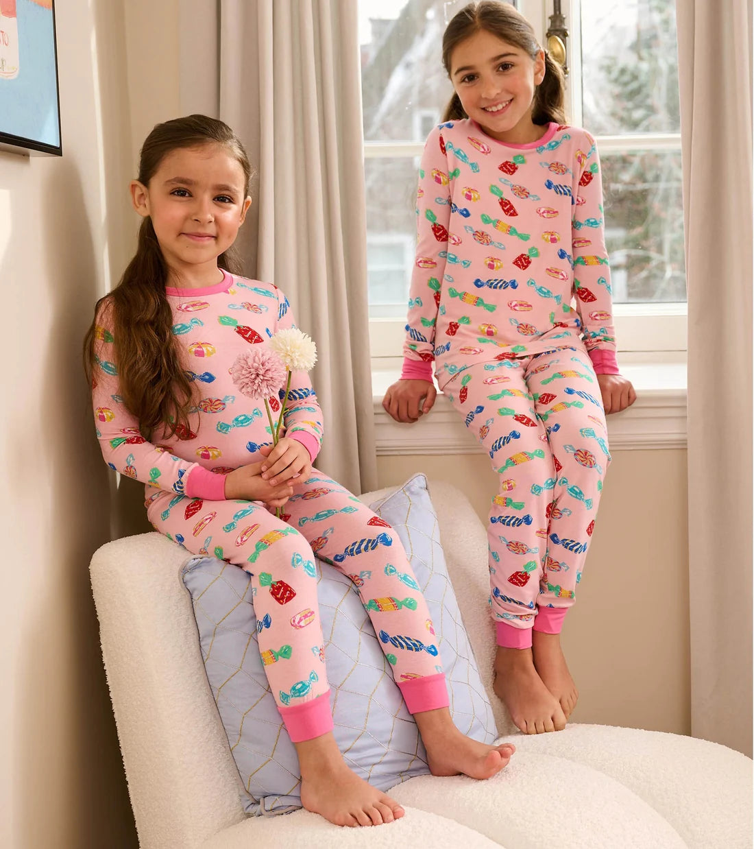 Two young girls wear matching Hatley Girls’ Delicious Candies Bamboo Pajama Sets as they pose in a sunlit room by the window—one sitting on a chair holding flowers, the other perched on the windowsill.