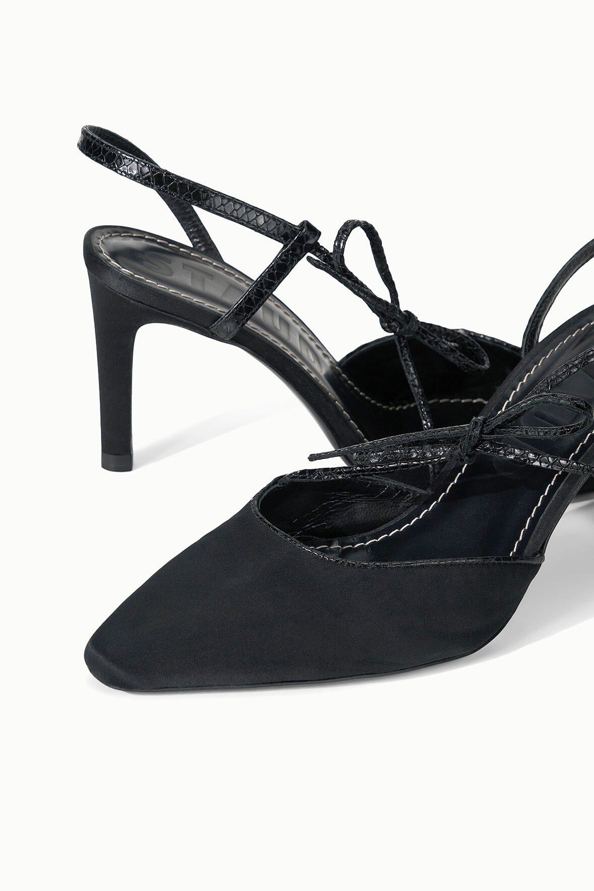 The Staud Inez Heel is a pair of black slingback heels with pointed toes, polished heels, and sophisticated crisscross strap detailing complemented by an elastic back strap for a secure fit—shown against a white background.